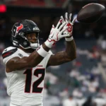 Nico Collins - Ein Footballspieler in einem weißen Trikot der Houston Texans mit der Nummer 12 fängt während eines Spiels einen Football, im Hintergrund sind verschwommene Stadionplätze zu sehen – dies unterstreicht den Moment, in dem Collins von den Texans gegen die Bengals ausfällt. Alt-Text wurde KI-generiert.
