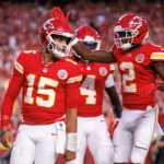 Patrick Mahomes - Drei Footballspieler der Kansas City Chiefs in roten Trikots stehen während eines Spiels auf dem Spielfeld; ein Spieler klopft einem anderen auf den Helm, während Mahomes trotz seiner Operation glänzt und mit seinem Team ein starkes Zeichen setzt. Alt-Text wurde KI-generiert.