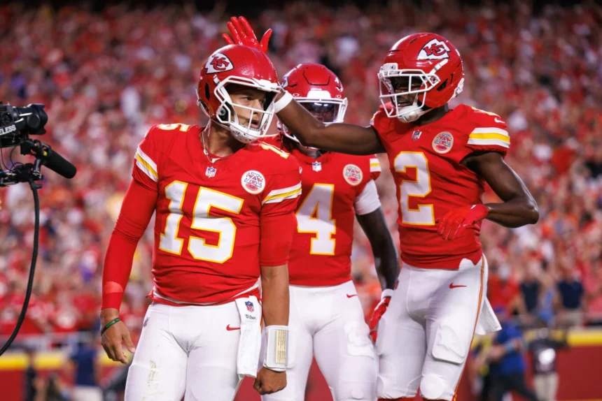 Patrick Mahomes - Drei Footballspieler der Kansas City Chiefs in roten Trikots stehen während eines Spiels auf dem Spielfeld; ein Spieler klopft einem anderen auf den Helm, während Mahomes trotz seiner Operation glänzt und mit seinem Team ein starkes Zeichen setzt. Alt-Text wurde KI-generiert.