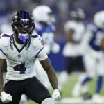 Zay Flowers - Ein Footballspieler der Baltimore Ravens in weißer Spielkleidung feiert während eines Spiels auf dem Spielfeld, während im Hintergrund Spieler der gegnerischen Mannschaft sowie seine eigenen Mannschaftskameraden zu sehen sind. Alt-Text wurde KI-generiert.