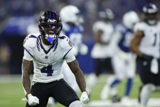 Zay Flowers - Ein Footballspieler der Baltimore Ravens in weißer Spielkleidung feiert während eines Spiels auf dem Spielfeld, während im Hintergrund Spieler der gegnerischen Mannschaft sowie seine eigenen Mannschaftskameraden zu sehen sind. Alt-Text wurde KI-generiert.
