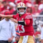 Brock Purdy - Ein Footballspieler im rot-goldenen Trikot der San Francisco 49ers macht sich bereit, während eines Spiels der zweiten NFL-Runde einen Football zu werfen, im Hintergrund eine Menge begeisterter Fans des Titelanwärters. Alt-Text wurde KI-generiert.