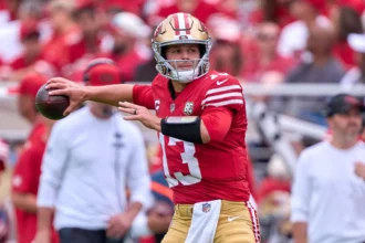 Brock Purdy - Ein Footballspieler im rot-goldenen Trikot der San Francisco 49ers macht sich bereit, während eines Spiels der zweiten NFL-Runde einen Football zu werfen, im Hintergrund eine Menge begeisterter Fans des Titelanwärters. Alt-Text wurde KI-generiert.