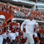 Die Footballspieler der Texas Longhorns und ihr Trainer Steve Sarkisian laufen auf das Spielfeld, während die Fans auf den Tribünen des Stadions jubeln. Alt-Text wurde KI-generiert.