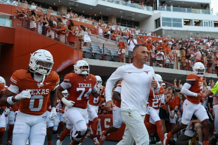 Die Footballspieler der Texas Longhorns und ihr Trainer Steve Sarkisian laufen auf das Spielfeld, während die Fans auf den Tribünen des Stadions jubeln. Alt-Text wurde KI-generiert.