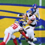 Jaxson Dart - Ein Football-Quarterback in weißer Spielkleidung wird von zwei Verteidigern in blauen und gelben Trikots auf einem Spielfeld mit einem großen gelb-blauen Logo zu Boden gebracht, während die Giants während des spannenden Spiels am Montag auf ihn zustürmen. Alt-Text wurde KI-generiert.