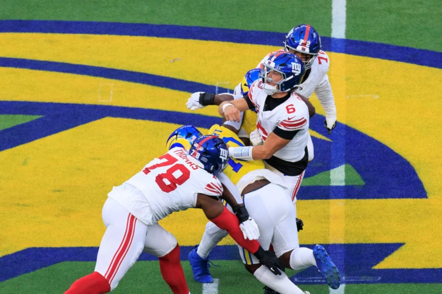 Jaxson Dart - Ein Football-Quarterback in weißer Spielkleidung wird von zwei Verteidigern in blauen und gelben Trikots auf einem Spielfeld mit einem großen gelb-blauen Logo zu Boden gebracht, während die Giants während des spannenden Spiels am Montag auf ihn zustürmen. Alt-Text wurde KI-generiert.
