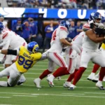 Aaron Donald - Footballspieler der New York Giants und der Los Angeles Rams sind mitten in einem Spielzug: Ein Verteidiger der Rams – Donald, der wieder im Einsatz ist – versucht, einen ballführenden Spieler der Giants zu tackeln, während Stafford die Rams entschlossen über das Feld führt. Alt-Text wurde KI-generiert.