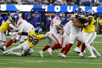 Aaron Donald - Footballspieler der New York Giants und der Los Angeles Rams sind mitten in einem Spielzug: Ein Verteidiger der Rams – Donald, der wieder im Einsatz ist – versucht, einen ballführenden Spieler der Giants zu tackeln, während Stafford die Rams entschlossen über das Feld führt. Alt-Text wurde KI-generiert.