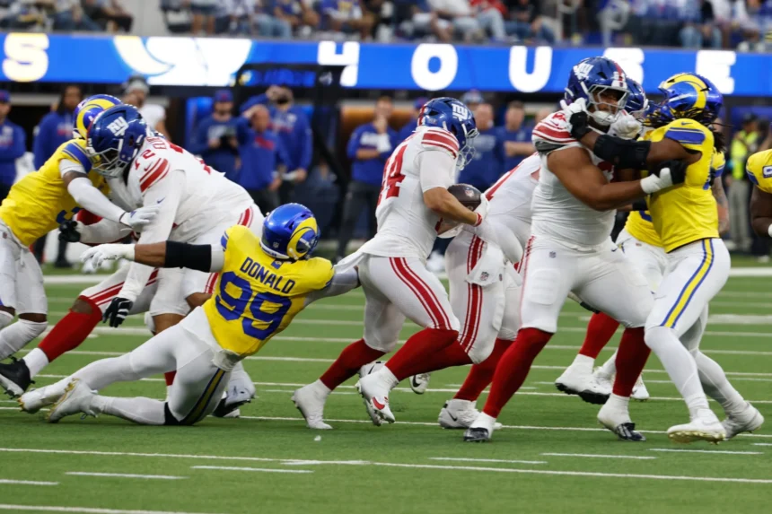 Aaron Donald - Footballspieler der New York Giants und der Los Angeles Rams sind mitten in einem Spielzug: Ein Verteidiger der Rams – Donald, der wieder im Einsatz ist – versucht, einen ballführenden Spieler der Giants zu tackeln, während Stafford die Rams entschlossen über das Feld führt. Alt-Text wurde KI-generiert.