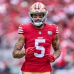 Mike Evans - Ein Footballspieler in einem roten Trikot der San Francisco 49ers mit der Nummer 5 auf dem Spielfeld, bekleidet mit Helm und Handschuhen, vor dem Hintergrund einer verschwommenen Stadionkulisse – Evans, der Starspieler der 49ers, bereitet sich auf das Spiel gegen die Cardinals vor. Alt-Text wurde KI-generiert.