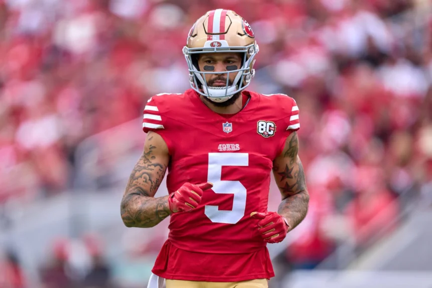 Mike Evans - Ein Footballspieler in einem roten Trikot der San Francisco 49ers mit der Nummer 5 auf dem Spielfeld, bekleidet mit Helm und Handschuhen, vor dem Hintergrund einer verschwommenen Stadionkulisse – Evans, der Starspieler der 49ers, bereitet sich auf das Spiel gegen die Cardinals vor. Alt-Text wurde KI-generiert.