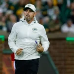 Matt LaFleur - Ein Mann in einer weißen Jacke und einer Kappe mit dem Logo der Green Bay Packers steht während eines Spiels auf einem Footballfeld und erinnert an Coach LaFleur, während er das Geschehen beobachtet. Alt-Text wurde KI-generiert.