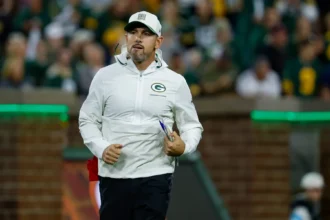 Matt LaFleur - Ein Mann in einer weißen Jacke und einer Kappe mit dem Logo der Green Bay Packers steht während eines Spiels auf einem Footballfeld und erinnert an Coach LaFleur, während er das Geschehen beobachtet. Alt-Text wurde KI-generiert.