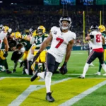 Bijan Robinson - Ein Footballspieler in weißer Spielkleidung rennt in die Endzone und sorgt für einen beeindruckenden Auftritt, während die Falcons bei einem Abendspiel gegen ihre Gegner antreten – im Hintergrund sind seine Teamkollegen und die Gegner zu sehen. Alt-Text wurde KI-generiert.