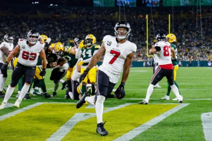 Bijan Robinson - Ein Footballspieler in weißer Spielkleidung rennt in die Endzone und sorgt für einen beeindruckenden Auftritt, während die Falcons bei einem Abendspiel gegen ihre Gegner antreten – im Hintergrund sind seine Teamkollegen und die Gegner zu sehen. Alt-Text wurde KI-generiert.