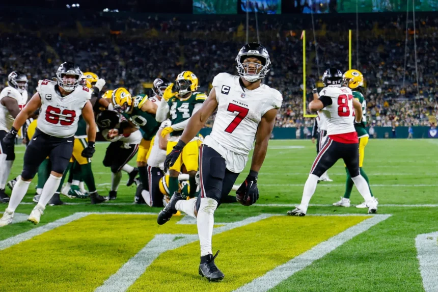 Bijan Robinson - Ein Footballspieler in weißer Spielkleidung rennt in die Endzone und sorgt für einen beeindruckenden Auftritt, während die Falcons bei einem Abendspiel gegen ihre Gegner antreten – im Hintergrund sind seine Teamkollegen und die Gegner zu sehen. Alt-Text wurde KI-generiert.