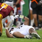 Dante Moore - Ein Footballspieler in weißer Kleidung liegt auf dem Spielfeld, während Moore aus Oregon, gekleidet in Rot und Gelb, während eines Spiels in der Nähe steht; im Hintergrund sind weitere Personen zu sehen. Alt-Text wurde KI-generiert.
