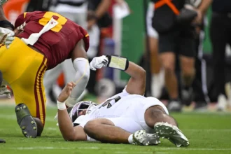 Dante Moore - Ein Footballspieler in weißer Kleidung liegt auf dem Spielfeld, während Moore aus Oregon, gekleidet in Rot und Gelb, während eines Spiels in der Nähe steht; im Hintergrund sind weitere Personen zu sehen. Alt-Text wurde KI-generiert.
