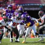 Ein Spieler der Minnesota Vikings – darunter Star-Receiver Justin Jefferson – läuft mit dem Football, während mehrere Verteidiger der Tampa Bay Buccaneers versuchen, ihn während eines NFL-Spiels zu tackeln. Alt-Text wurde KI-generiert.