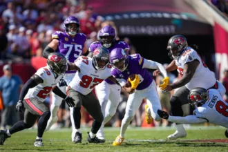 Ein Spieler der Minnesota Vikings – darunter Star-Receiver Justin Jefferson – läuft mit dem Football, während mehrere Verteidiger der Tampa Bay Buccaneers versuchen, ihn während eines NFL-Spiels zu tackeln. Alt-Text wurde KI-generiert.