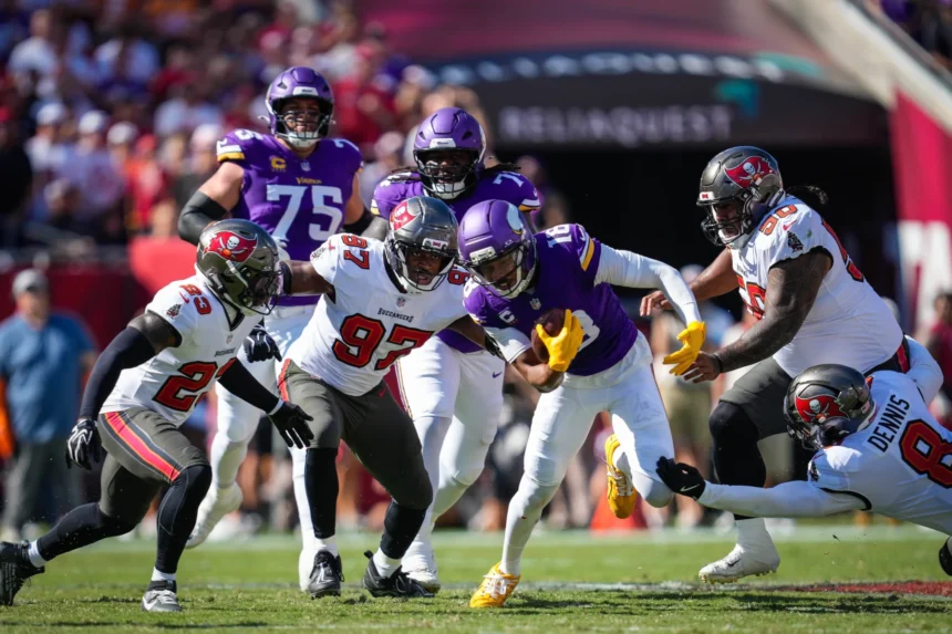 Ein Spieler der Minnesota Vikings – darunter Star-Receiver Justin Jefferson – läuft mit dem Football, während mehrere Verteidiger der Tampa Bay Buccaneers versuchen, ihn während eines NFL-Spiels zu tackeln. Alt-Text wurde KI-generiert.