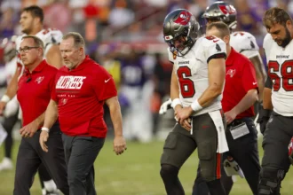 Baker Mayfield - Ein Footballspieler im Trikot der Buccaneers verlässt während eines Spiels zusammen mit Trainern und Betreuern das Spielfeld, hält sich den Arm und scheint verletzt zu sein – Mayfield fällt wegen einer Daumenverletzung mindestens für den Rest des Spiels aus. Alt-Text wurde KI-generiert.