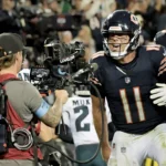 Case Keenum - Ein Footballspieler in einem dunklen Trikot mit der Nummer 11 schreit während eines Spiels auf dem Spielfeld und gestikuliert in Richtung eines Fernsehkameramanns – und sendet damit eine klare Botschaft: Kein Caleb, kein Problem. Im Hintergrund sind weitere Spieler und Zuschauer zu sehen. Alt-Text wurde KI-generiert.