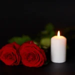 Zwei rote Southern-Nazarene-Rosen und eine brennende weiße Kerze auf dunklem Hintergrund.