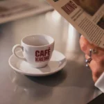 Eine Tasse Café Krewe-Kaffee steht auf einer Untertasse neben einer Person, die im „Buccaneers Café“ eine Zeitung liest.