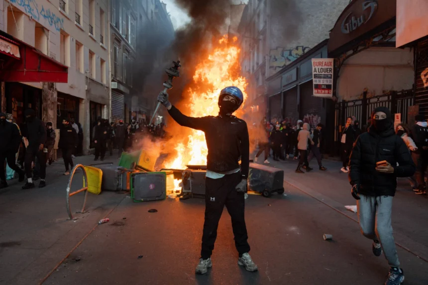 Eine maskierte Person mit Helm steht auf einer Straße in Paris und hält eine Laterne über einen brennenden Trümmerhaufen, während Menschen vorbeigehen.