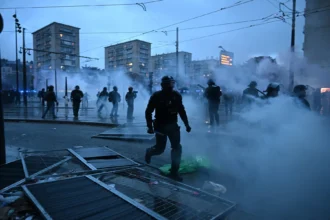 Polizisten in Schutzausrüstung stehen auf einer verrauchten Straße vor dem geschlossenen NFL House in Paris; im Vordergrund liegen umgestürzte Metallbarrikaden.