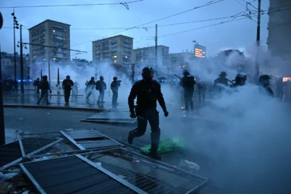 Polizisten in Schutzausrüstung stehen auf einer verrauchten Straße vor dem geschlossenen NFL House in Paris; im Vordergrund liegen umgestürzte Metallbarrikaden.