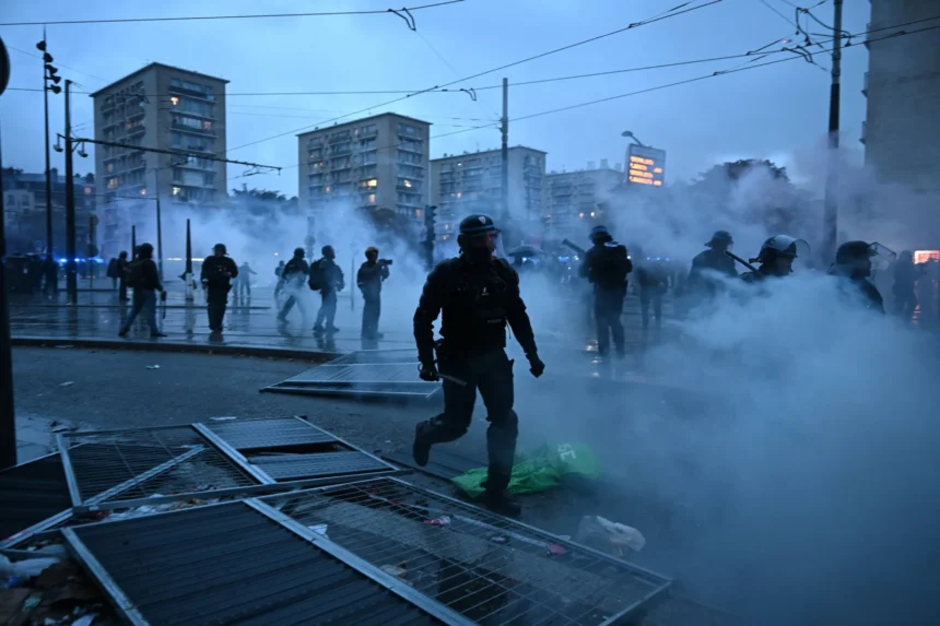 Polizisten in Schutzausrüstung stehen auf einer verrauchten Straße vor dem geschlossenen NFL House in Paris; im Vordergrund liegen umgestürzte Metallbarrikaden.