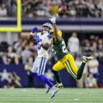 Ein Spieler der Green Bay Packers im weißen Trikot und ein Spieler der Cowboys im gelb-grünen Trikot springen in die Luft, um einen Ball zu fangen.