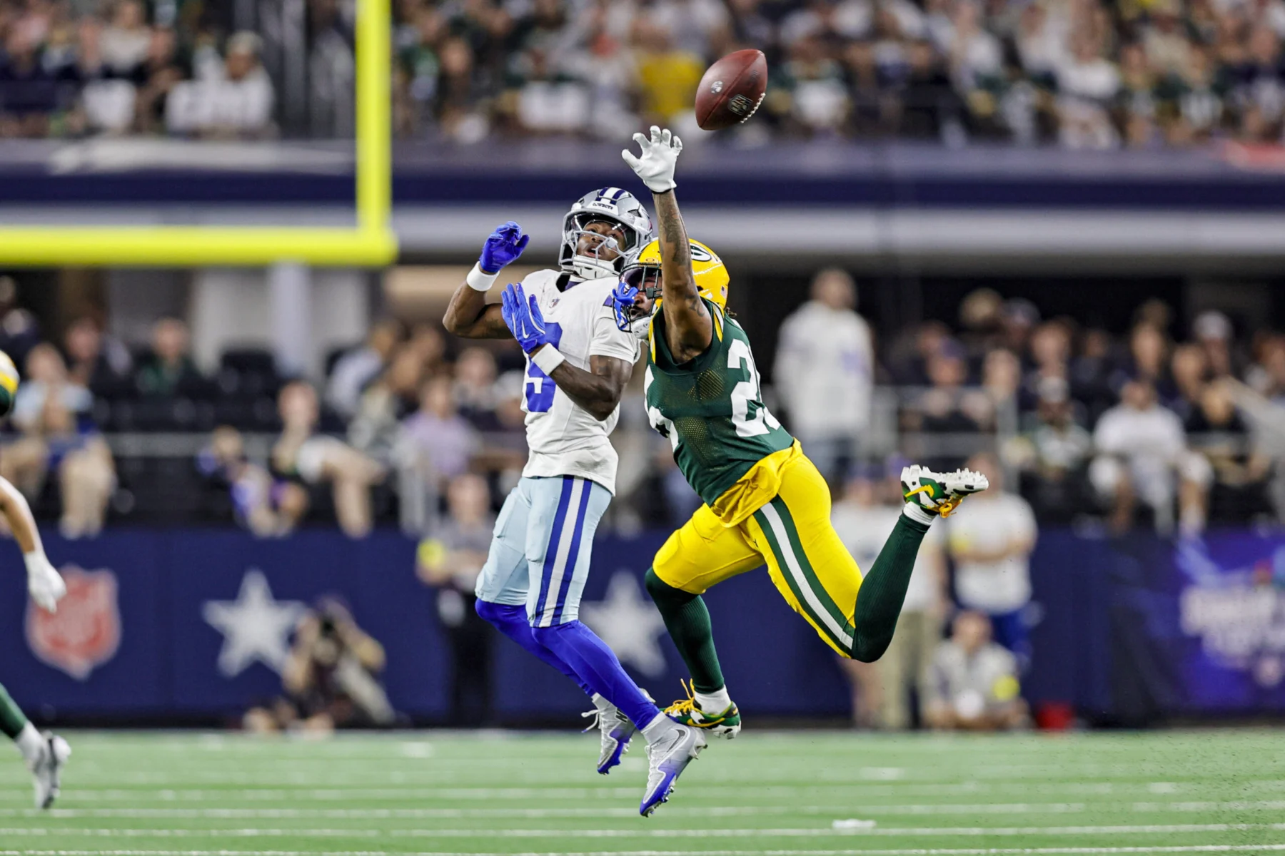 Ein Spieler der Green Bay Packers im weißen Trikot und ein Spieler der Cowboys im gelb-grünen Trikot springen in die Luft, um einen Ball zu fangen.