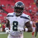 Jadarian Price - Ein Footballspieler in Trikot und Helm der Seattle Seahawks steht auf dem Spielfeld, während die Seahawks Price wegen seiner Leistung in einem roten Stadion mit anderen Spielern im Hintergrund aufstellen. Alt-Text wurde KI-generiert.