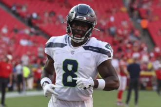 Jadarian Price - Ein Footballspieler in Trikot und Helm der Seattle Seahawks steht auf dem Spielfeld, während die Seahawks Price wegen seiner Leistung in einem roten Stadion mit anderen Spielern im Hintergrund aufstellen. Alt-Text wurde KI-generiert.
