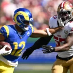 Ein Footballspieler in einem blau-gelben Trikot, das an die Ära der Rams unter Coach McVay erinnert, wehrt während eines Spiels mit ausgestrecktem Arm einen Verteidiger in Weiß und Rot ab, während er den Ball trägt. Alt-Text wurde KI-generiert.