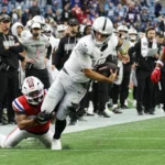 Fernando Mendoza - Ein Quarterback der Raiders in weißem Trikot läuft mit dem Football, während er von einem Spieler der Patriots in rotem Trikot zu Boden gebracht wird – Mendoza gibt dabei seine NFL-Performance zum Besten. Alt-Text wurde KI-generiert.