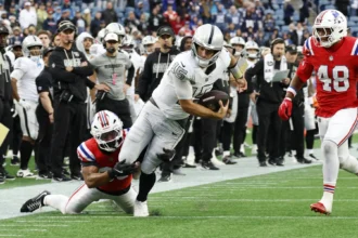 Fernando Mendoza - Ein Quarterback der Raiders in weißem Trikot läuft mit dem Football, während er von einem Spieler der Patriots in rotem Trikot zu Boden gebracht wird – Mendoza gibt dabei seine NFL-Performance zum Besten. Alt-Text wurde KI-generiert.