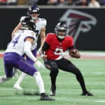 Michael Penix Jr., Quarterback der Atlanta Falcons, hält einen Football in der Hand, während er von zwei Verteidigern der Baltimore Ravens verfolgt wird – dies geschah während eines Spiels, bei dem sich Penix eine Rippenverletzung zugezogen hatte. Alt-Text wurde KI-generiert.