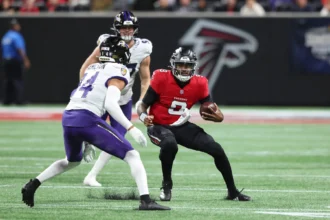 Michael Penix Jr., Quarterback der Atlanta Falcons, hält einen Football in der Hand, während er von zwei Verteidigern der Baltimore Ravens verfolgt wird – dies geschah während eines Spiels, bei dem sich Penix eine Rippenverletzung zugezogen hatte. Alt-Text wurde KI-generiert.