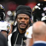 Lamar Jackson trägt während eines Spiels einen schwarzen Kapuzenpullover und ein Durag, umgeben von seinen Teamkollegen der Baltimore Ravens, die Helme tragen.