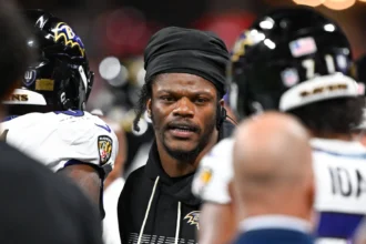 Lamar Jackson trägt während eines Spiels einen schwarzen Kapuzenpullover und ein Durag, umgeben von seinen Teamkollegen der Baltimore Ravens, die Helme tragen.