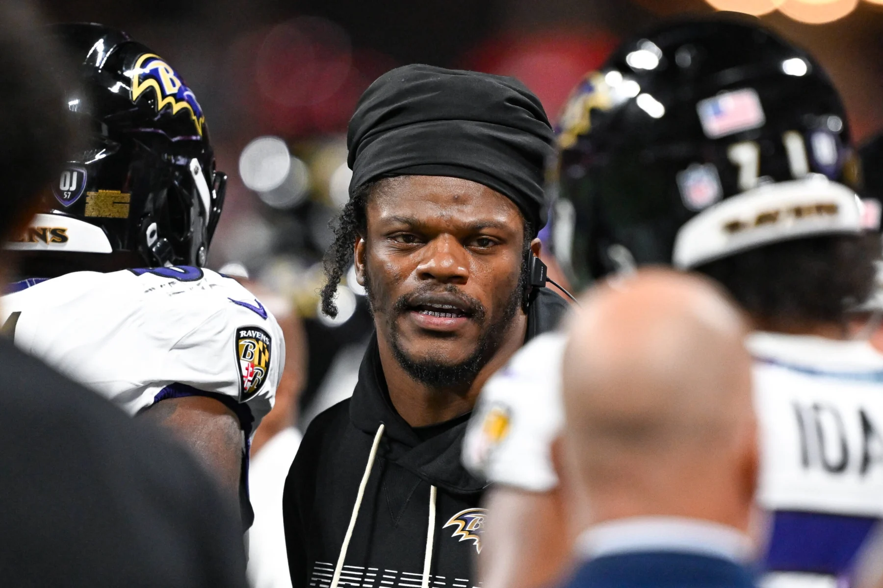 Lamar Jackson trägt während eines Spiels einen schwarzen Kapuzenpullover und ein Durag, umgeben von seinen Teamkollegen der Baltimore Ravens, die Helme tragen.