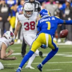 Chad Ryland - Ein Spieler der Arizona Cardinals in weißer Spielkleidung schießt den Ball, während ein Teamkollege kniet und ein Spieler in blau-gelber Spielkleidung versucht, den Schuss zu blocken.