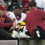 Terry McLaurin, ein Spieler der Washington Commanders, sitzt auf einem motorisierten Golfwagen zwischen zwei Mitarbeitern des Teams in kastanienbraunen Jacken.