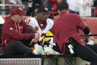 Terry McLaurin, ein Spieler der Washington Commanders, sitzt auf einem motorisierten Golfwagen zwischen zwei Mitarbeitern des Teams in kastanienbraunen Jacken.