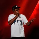 Jay-Z tritt beim Super Bowl LXI auf der Bühne auf und trägt eine Sonnenbrille, eine schwarze Baseballkappe sowie ein weißes T-Shirt mit einem Beatles-Motiv.